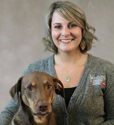 Emily Ferber smiling in front of a grey backdrop hugging a brown dog