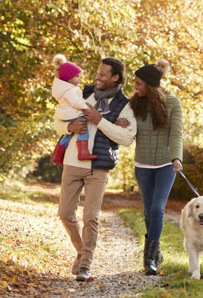 A husband and wife walking in a park with their golden retriever as the dad and mom are smiling and holding (dad) their toddler.