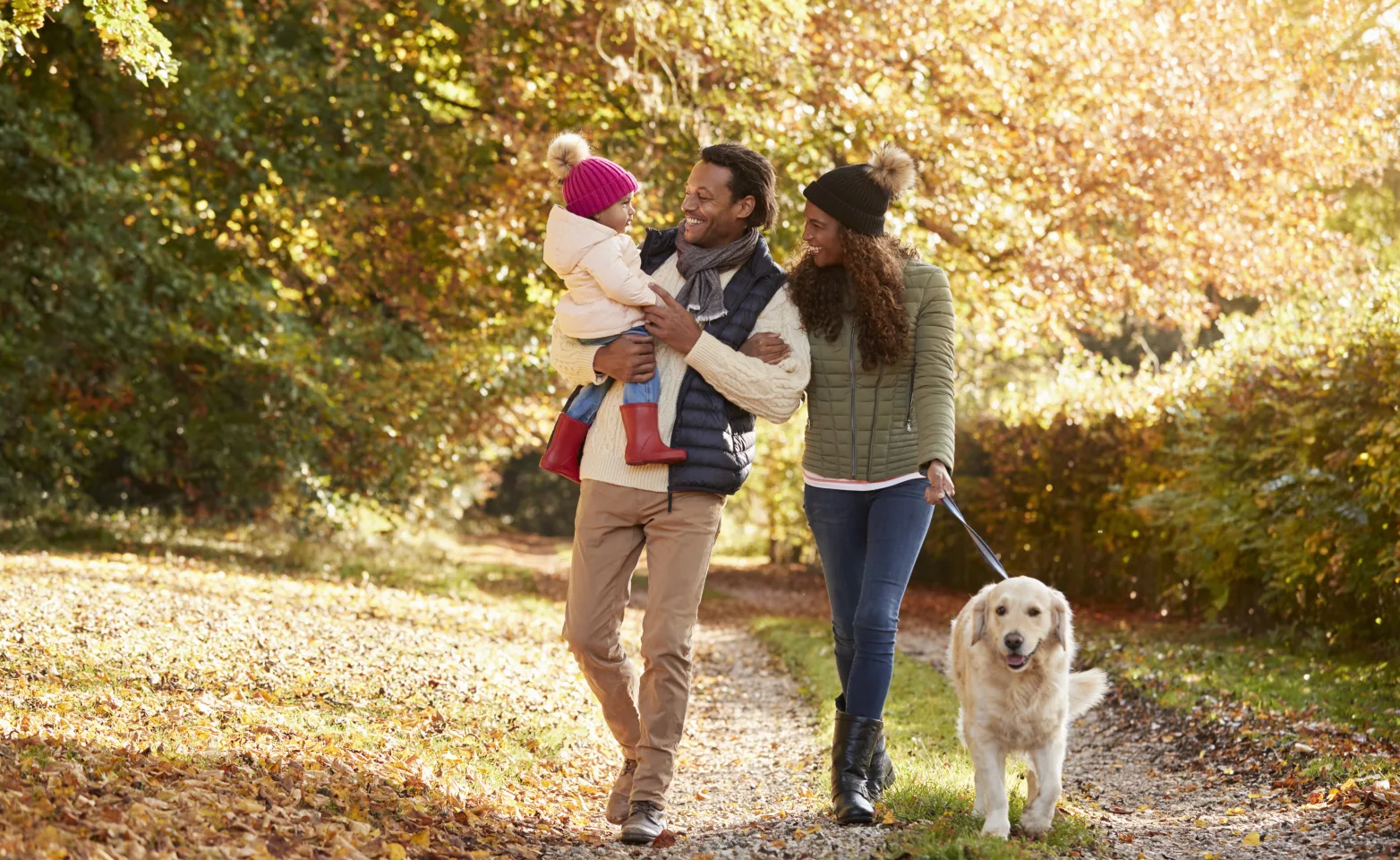 A husband and wife walking in a park with their golden retriever as the dad and mom are smiling and holding (dad) their toddler.