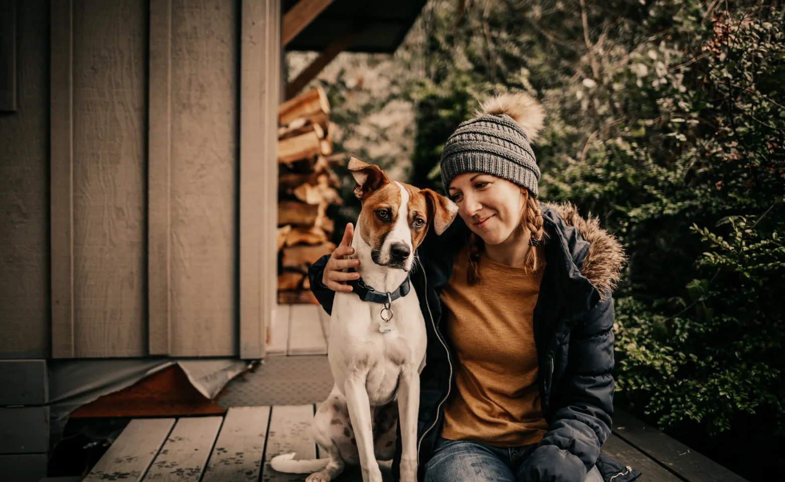 dog and woman sitting on a wooden deck with chopped wood and shrubbery in the background