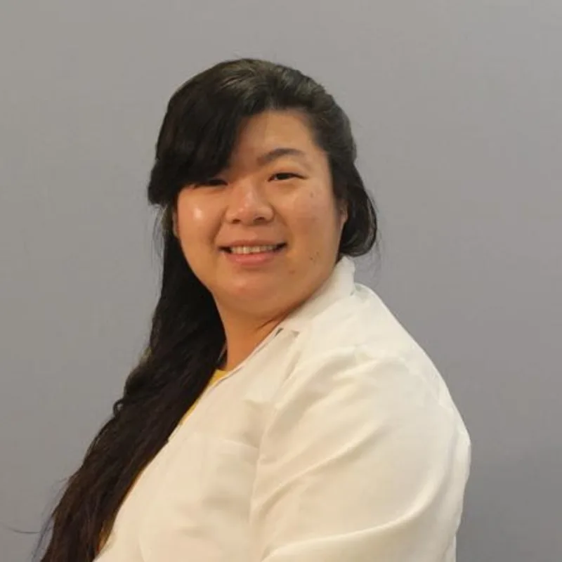 Rosa Yip smiling in front of a grey wall wearing a white lab coat Rosa Yip smiling in front of a grey wall wearing a white lab coat