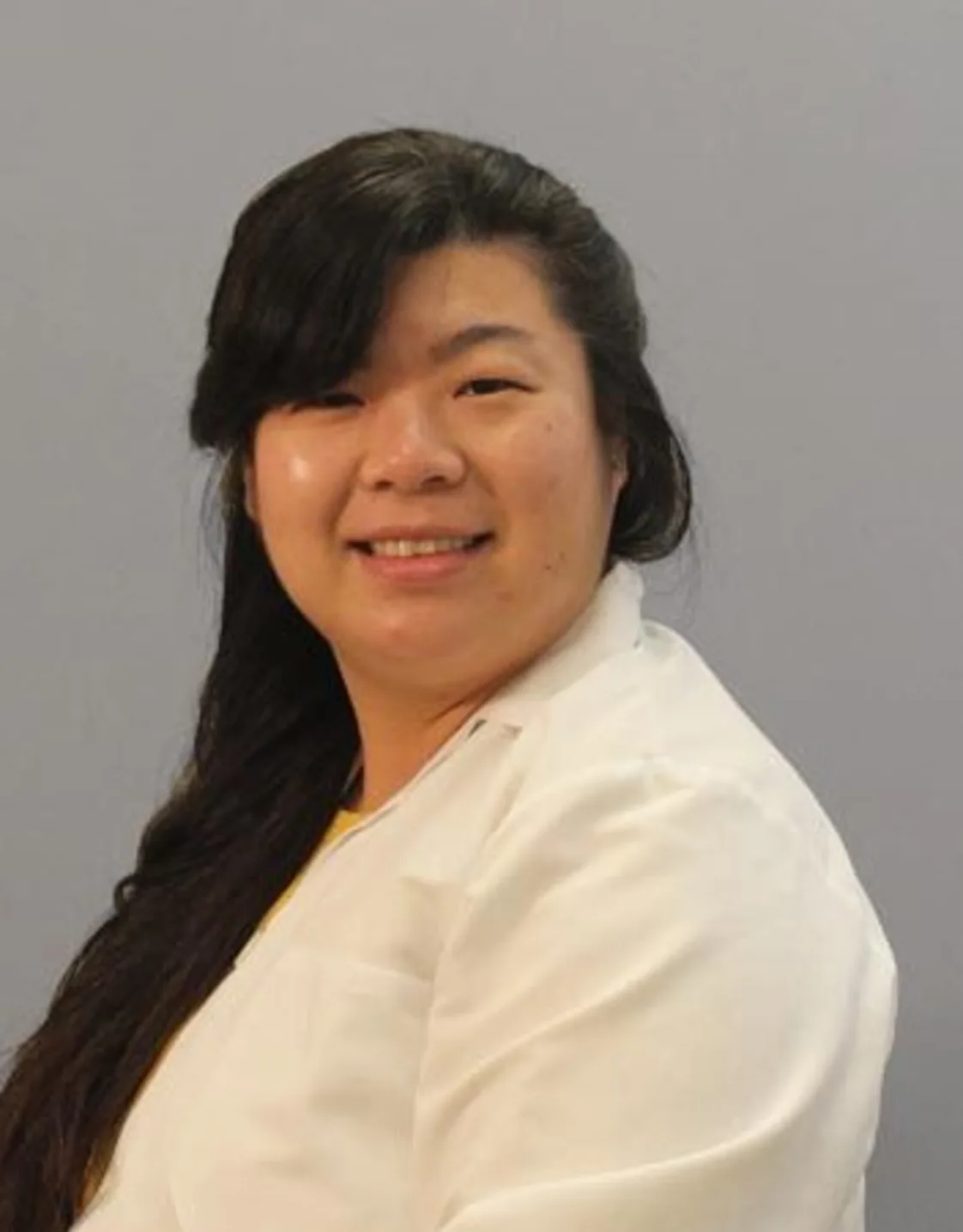 Rosa Yip smiling in front of a grey wall wearing a white lab coat Rosa Yip smiling in front of a grey wall wearing a white lab coat