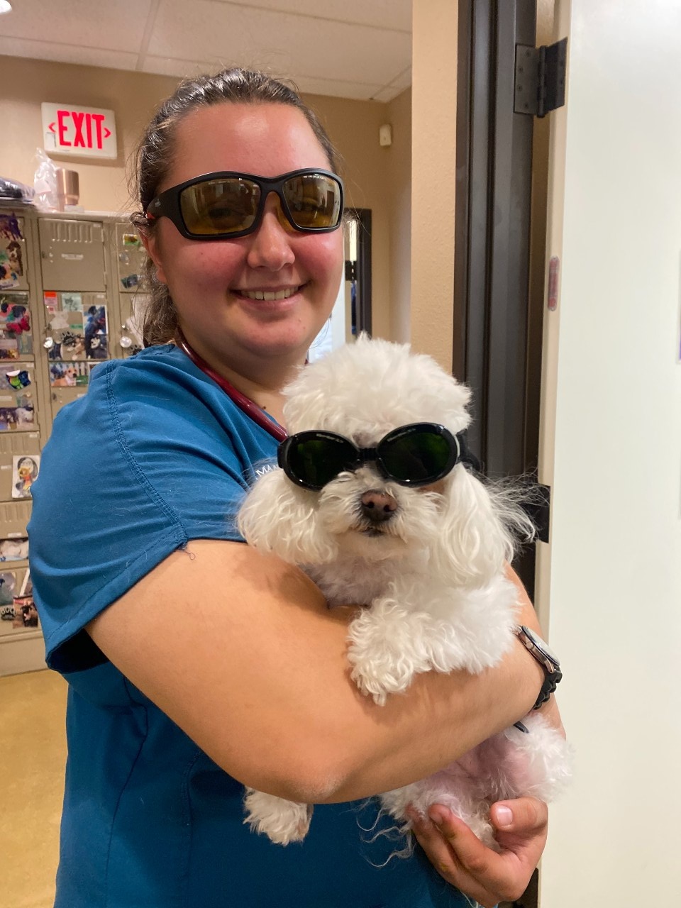Woman in blue scrubs holding white fluffy dog while they both wear sunglasses.