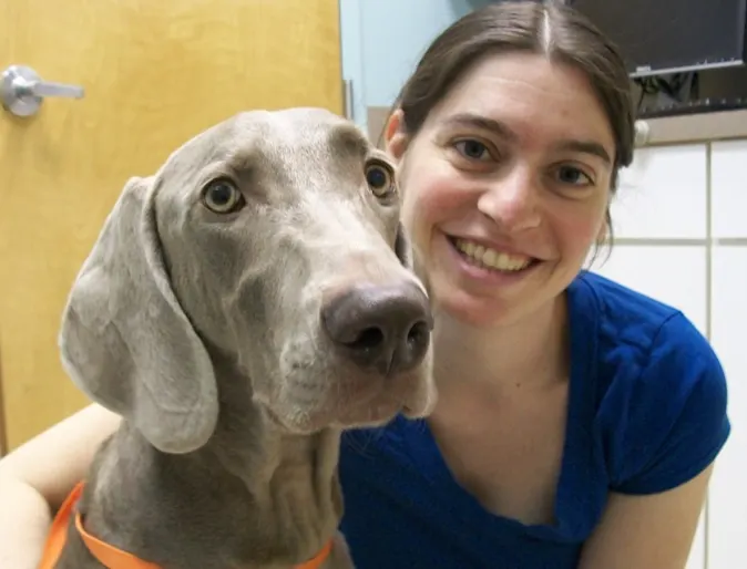 Woman in a blue shirt smiling next to Hobbes, a Weimaraner Woman in a blue shirt smiling next to Hobbes, a Weimaraner