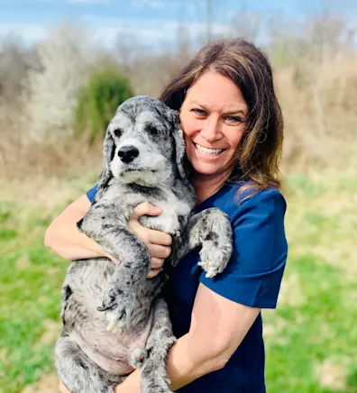 Lynette holding onto a small dog while smiling in a grassy plain. Lynette holding onto a small dog while smiling in a grassy plain.