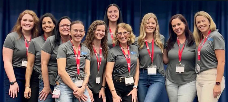 A group of ten women in matching gray shirts and red lanyards pose together at a conference