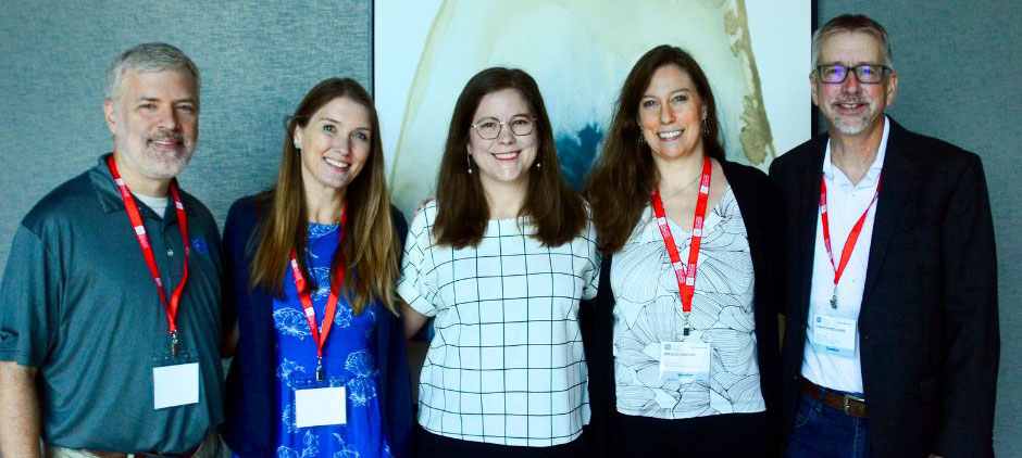 Five teammates wearing red lanyards pose together in front of a presentation screen