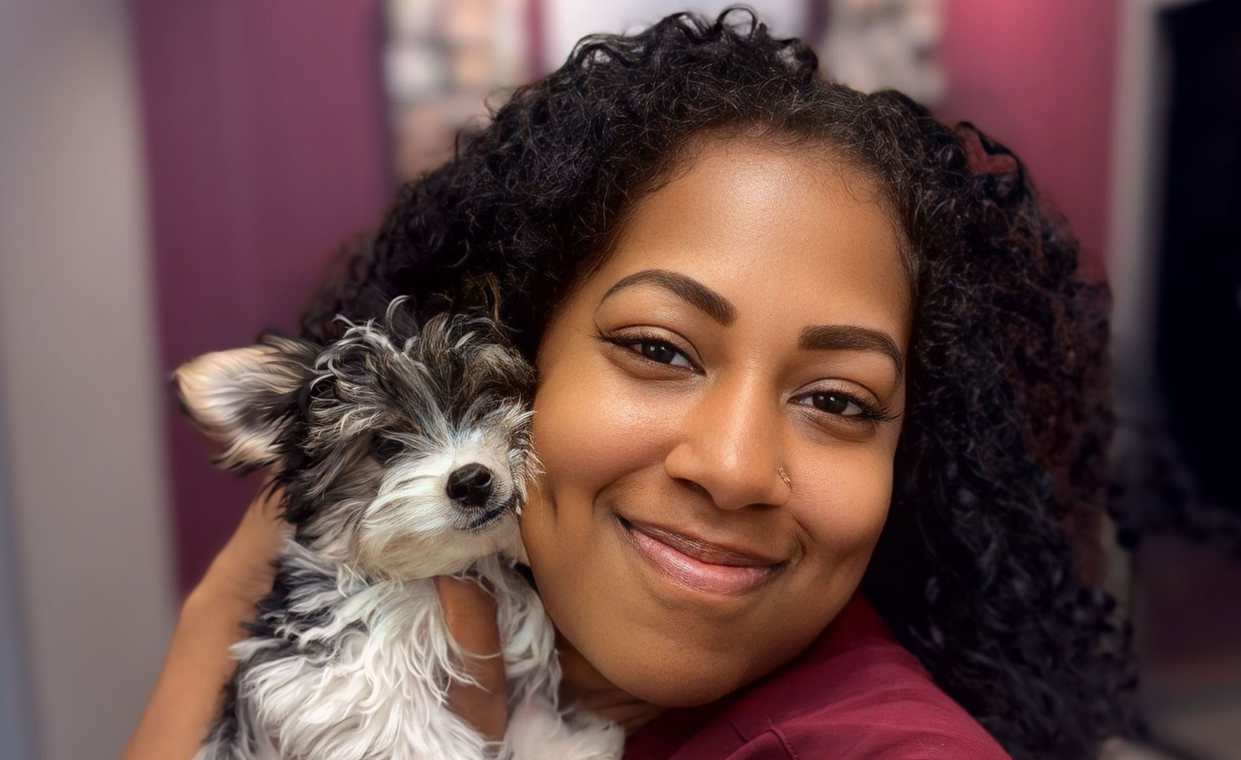 Close-up of a woman with curly dark hair in a maroon scrub top smiling with a small gray-and-white terrier puppy