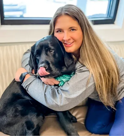 Rachel McIntyre smiling hugging a Black Lab sitting on waiting bench Rachel McIntyre smiling hugging a Black Lab sitting on waiting bench
