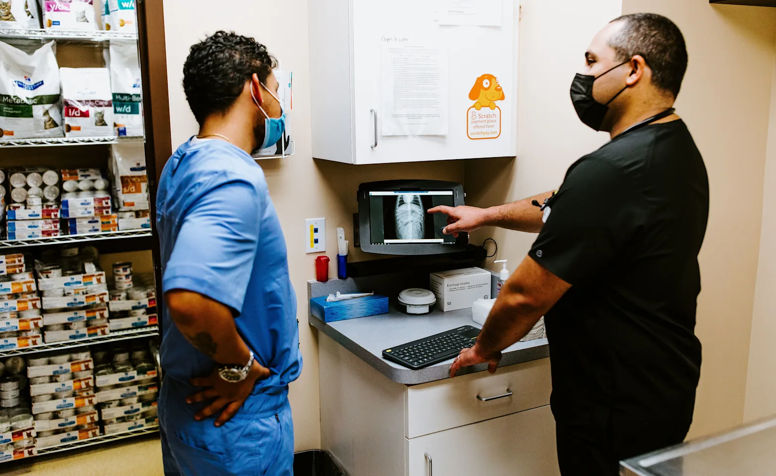 Two healthcare professionals discuss an X-ray image on a monitor in a medical office. Both are wearing masks and scrubs. Shelves stocked with supplies are visible in the background. Two healthcare professionals discuss an X-ray image on a monitor in a medical office. Both are wearing masks and scrubs. Shelves stocked with supplies are visible in the background.