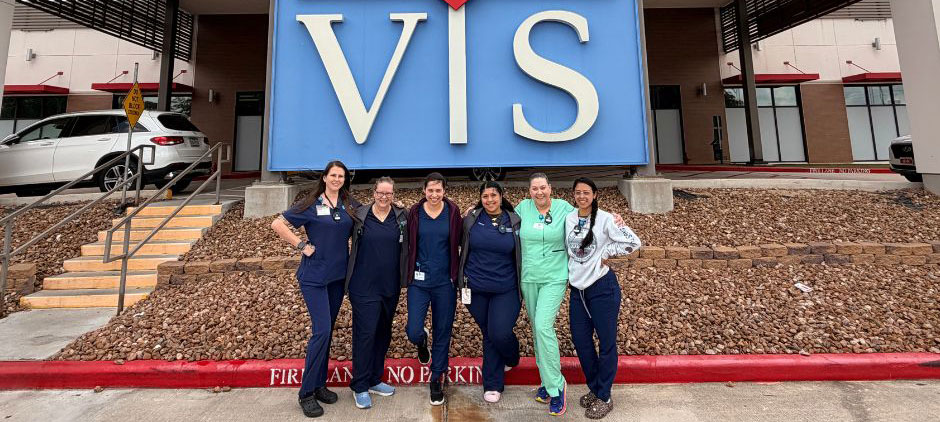 Six veterinary staff members in scrubs pose together in front of a large outdoor sign