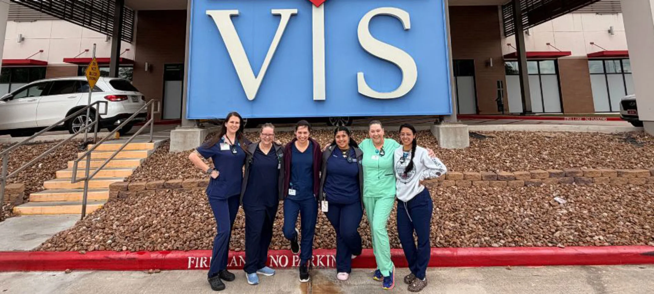Six veterinary staff members in scrubs pose together in front of a large outdoor sign Six veterinary staff members in scrubs pose together in front of a large outdoor sign