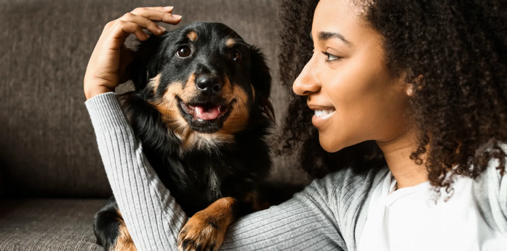 A woman sitting in front of a couch with her arm around a black and brown dog. 