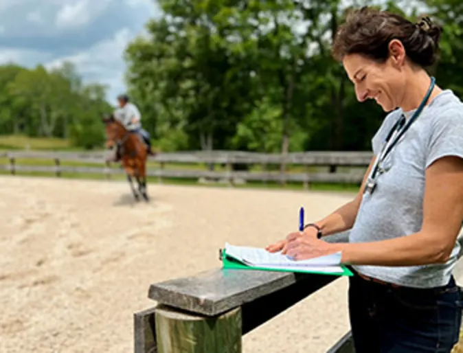 Woman standing taking notes while a horse and rider are in the background