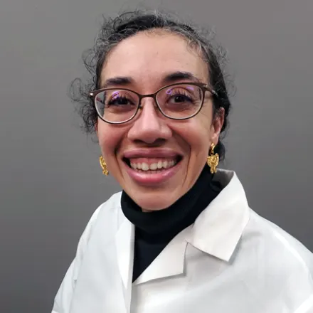 Terra Martin smiling standing in front of a grey wall wearing a white lab coat