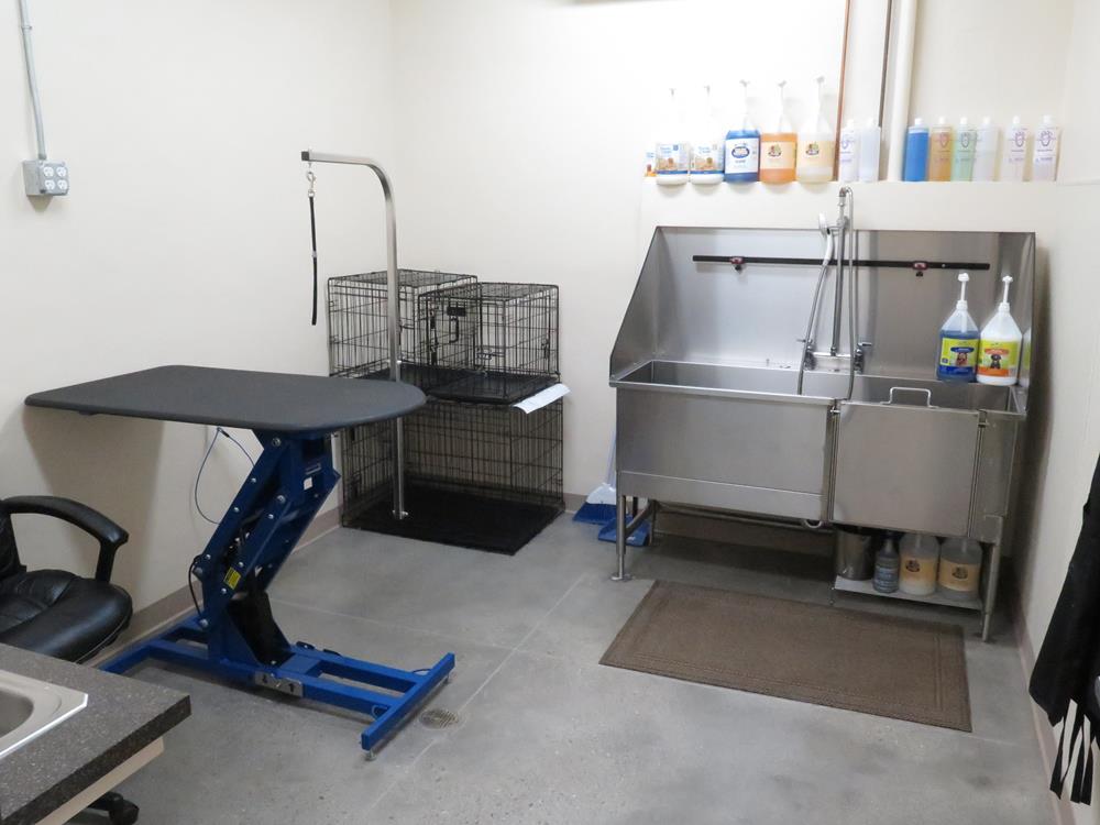 The grooming area room show a side table sticking out of the wall, a large shower sink for animals, shampoos and a cage in the corner of the room. 