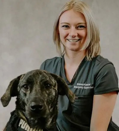 Brianna Koester smiling in front of a grey backdrop kneeling next to a large black and brown dog