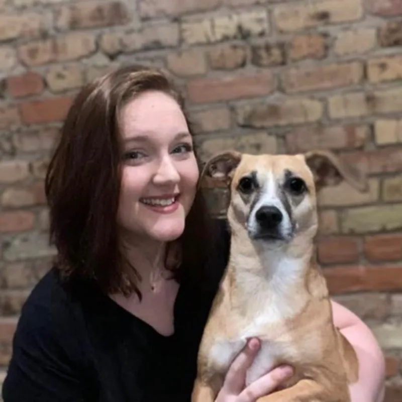 Kacie standing in front of a brick wall holding a small light brown and white dog Kacie standing in front of a brick wall holding a small light brown and white dog