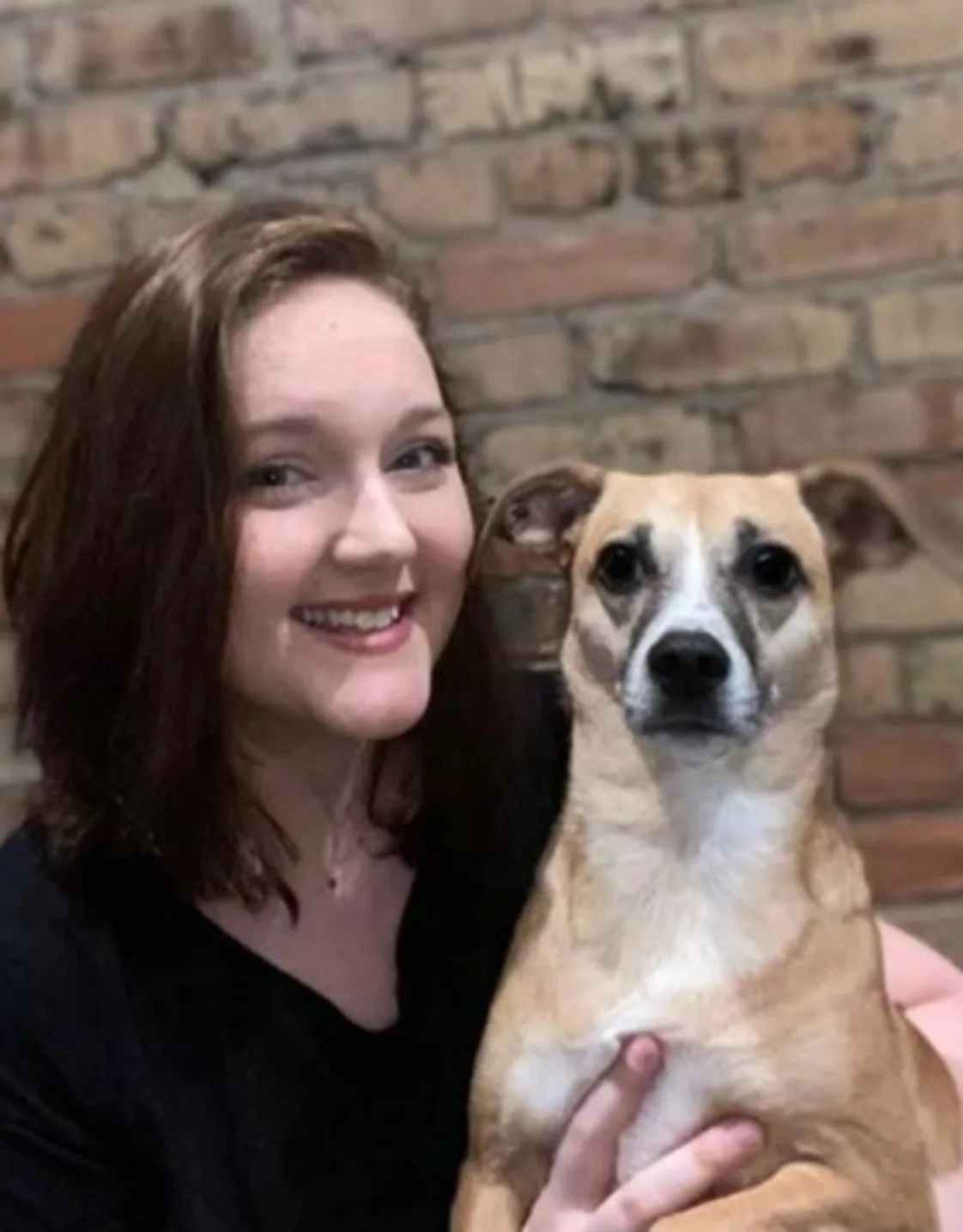 Kacie standing in front of a brick wall holding a small light brown and white dog Kacie standing in front of a brick wall holding a small light brown and white dog
