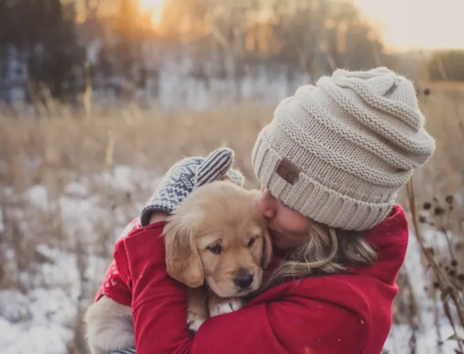 Woman holding a puppy in her arms outside in a field during winter time. She is wearing a pink - reddish coat with mittens and beanie. Woman holding a puppy in her arms outside in a field during winter time. She is wearing a pink - reddish coat with mittens and beanie.