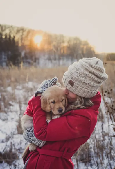 Woman holding a puppy in her arms outside in a field during winter time.  She is wearing a pink - reddish coat with mittens and beanie. Woman holding a puppy in her arms outside in a field during winter time.  She is wearing a pink - reddish coat with mittens and beanie.