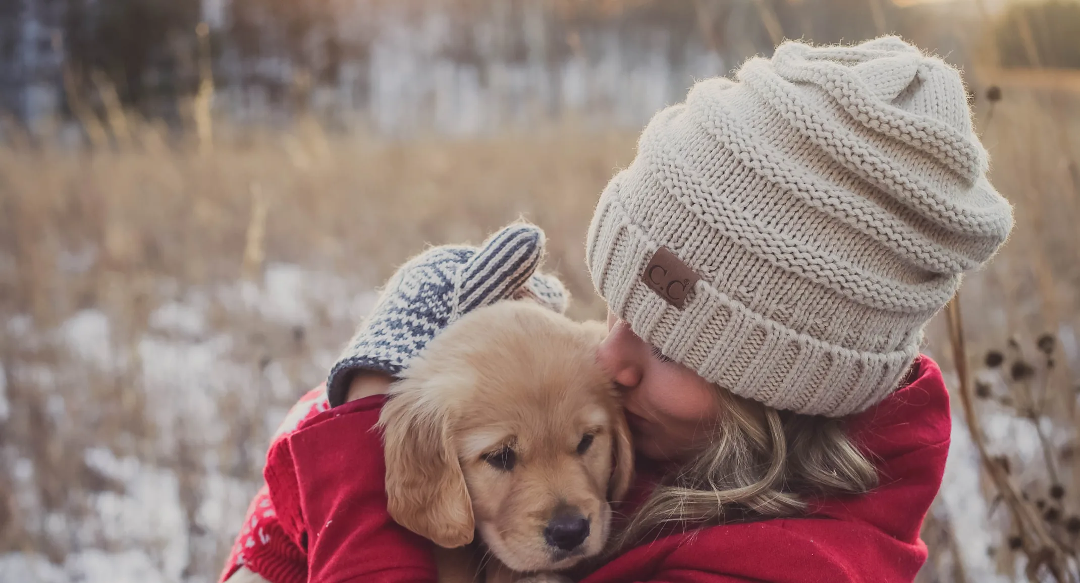 Woman holding a puppy in her arms outside in a field during winter time.  She is wearing a pink - reddish coat with mittens and beanie. Woman holding a puppy in her arms outside in a field during winter time.  She is wearing a pink - reddish coat with mittens and beanie.