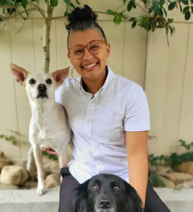 Ashley's staff photo from Valley Animal Hospital & Pet Resort where she is at a park holding two small dogs in each arm.