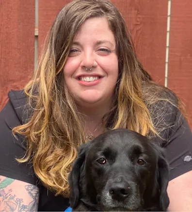 Jennifer in front of a brick wall kneeling down next to a black dog with a pink leash Jennifer in front of a brick wall kneeling down next to a black dog with a pink leash