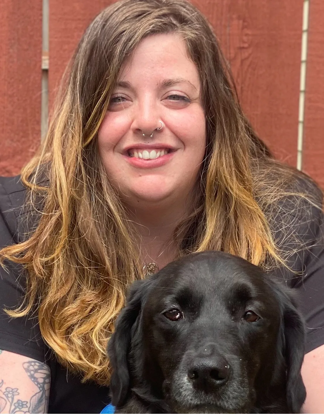 Jennifer in front of a brick wall kneeling down next to a black dog with a pink leash Jennifer in front of a brick wall kneeling down next to a black dog with a pink leash