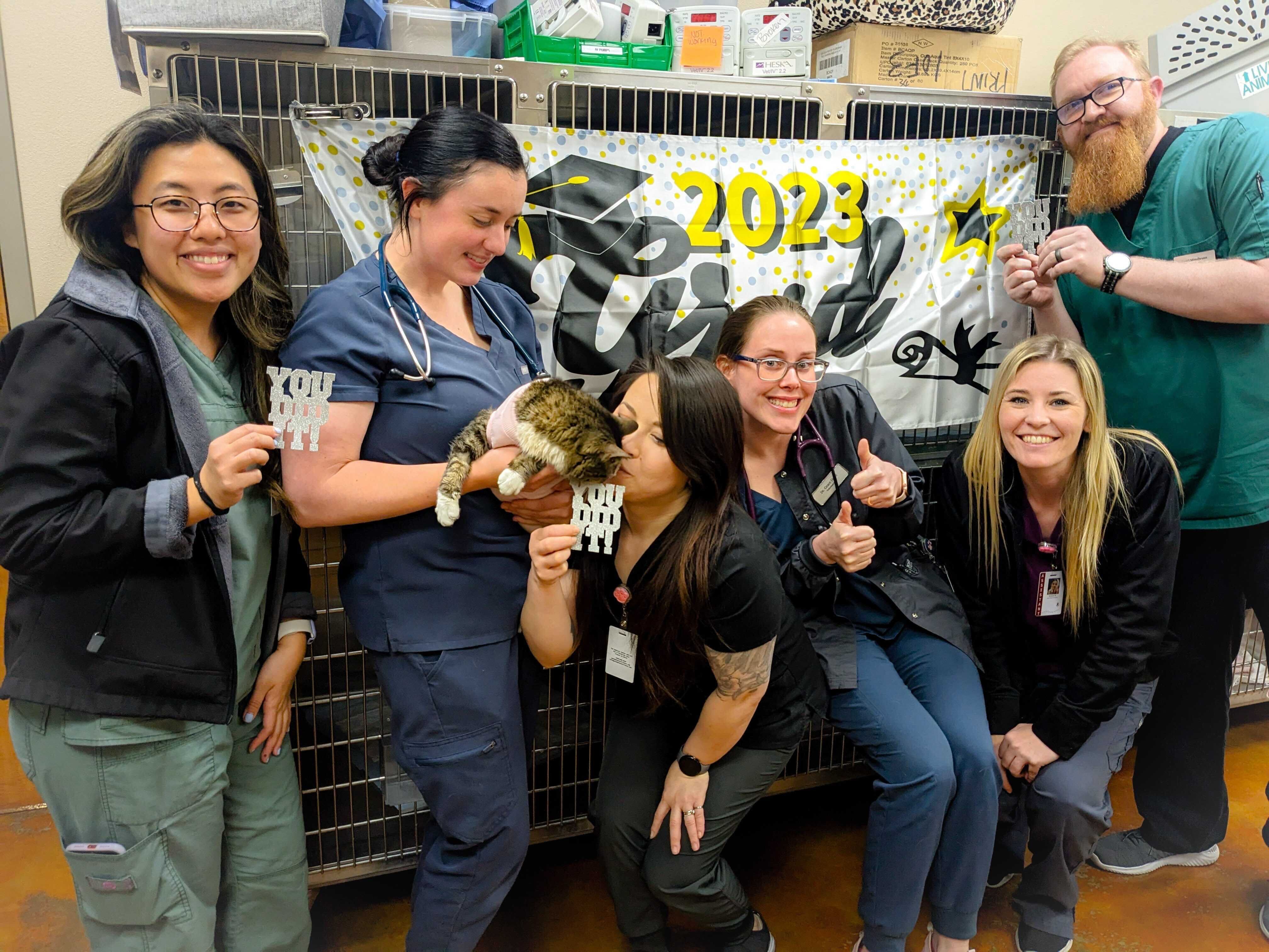 Employees posing with a cat and a sign that reads "2023 Grad"