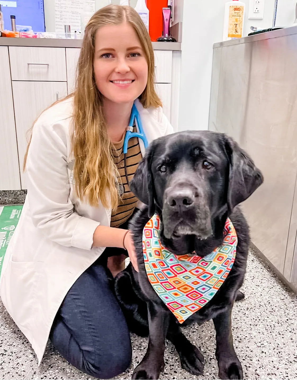 Dr. Elise Mauer smiling kneeling down next to a black dog wearing a bandana Dr. Elise Mauer smiling kneeling down next to a black dog wearing a bandana