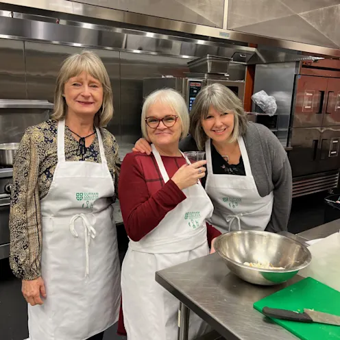 Sandra McBride smiling in a kitchen cooking with two friends. Sandra McBride smiling in a kitchen cooking with two friends.