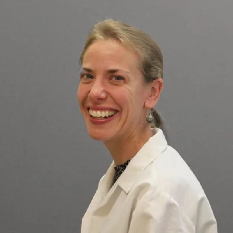 Suzanne Taylor smiling in front of a grey wall wearing a white lab coat Suzanne Taylor smiling in front of a grey wall wearing a white lab coat