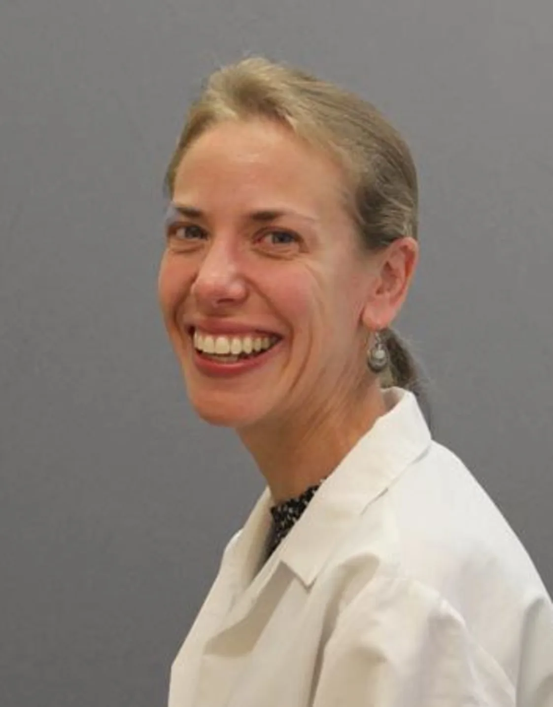 Suzanne Taylor smiling in front of a grey wall wearing a white lab coat Suzanne Taylor smiling in front of a grey wall wearing a white lab coat