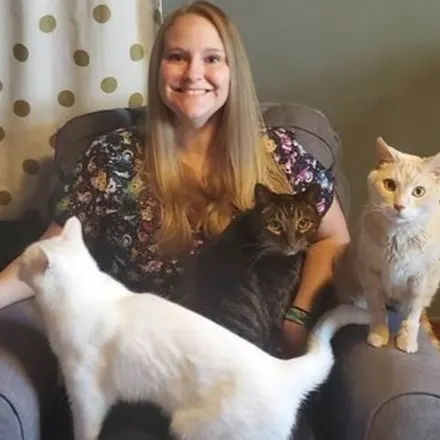 Staff photo from Niles Veterinary Clinic of Kourtney sitting on a blue couch with her two cats and little dog on her lap and arm chair, smiling.