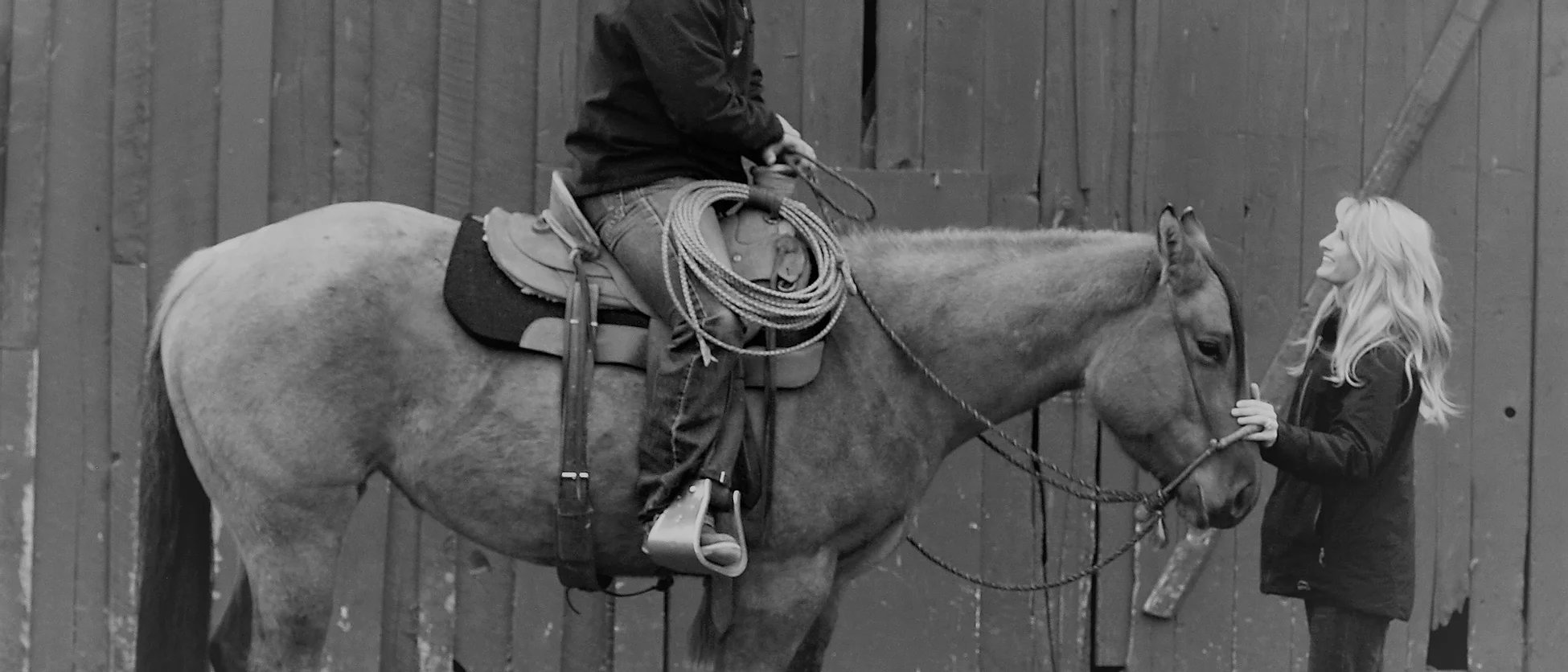 Black and white image of someone sitting on a horse while there is a woman in front.