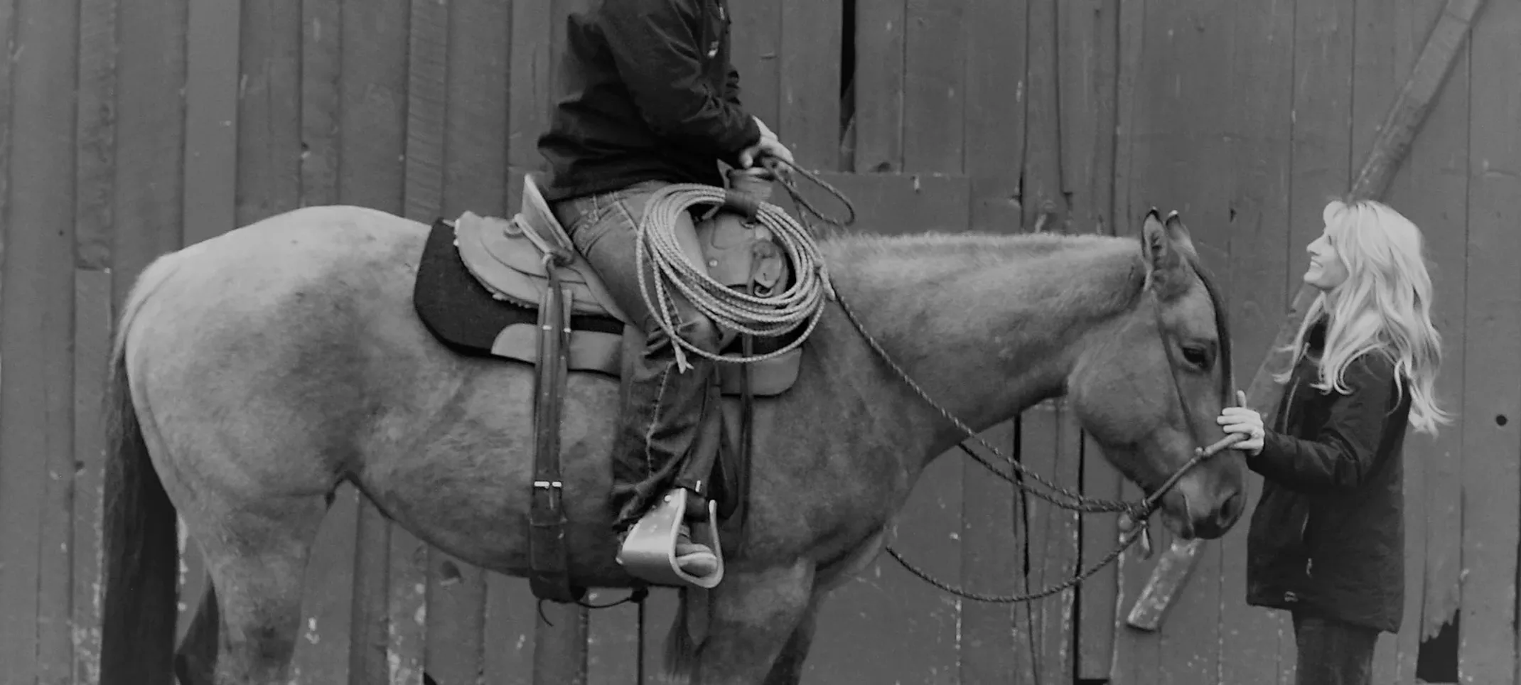 Black and white image of someone sitting on a horse while there is a woman in front.