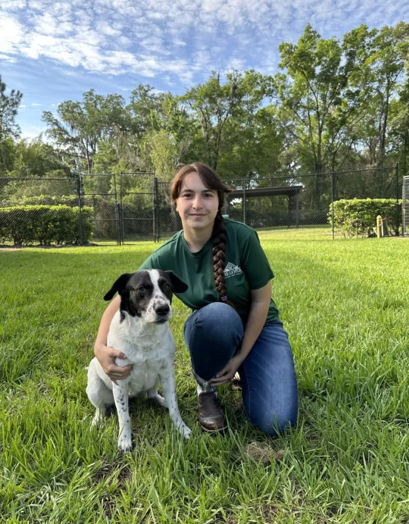 Shiann kneeling next to a medium sized dog with a background of green grass, tall trees, and blue skies