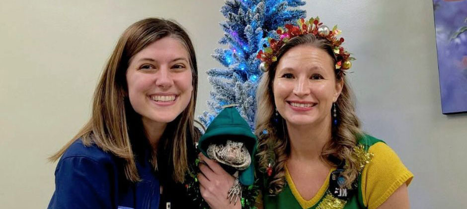 Two smiling women in festive attire pose with a bearded dragon dressed in a tiny holiday costume near a decorated tree
