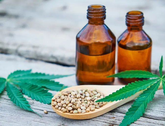 Two bottle of CDB Marijuana brown bottles next to each other next to Marijauna leaves, seeds on wooden table. 