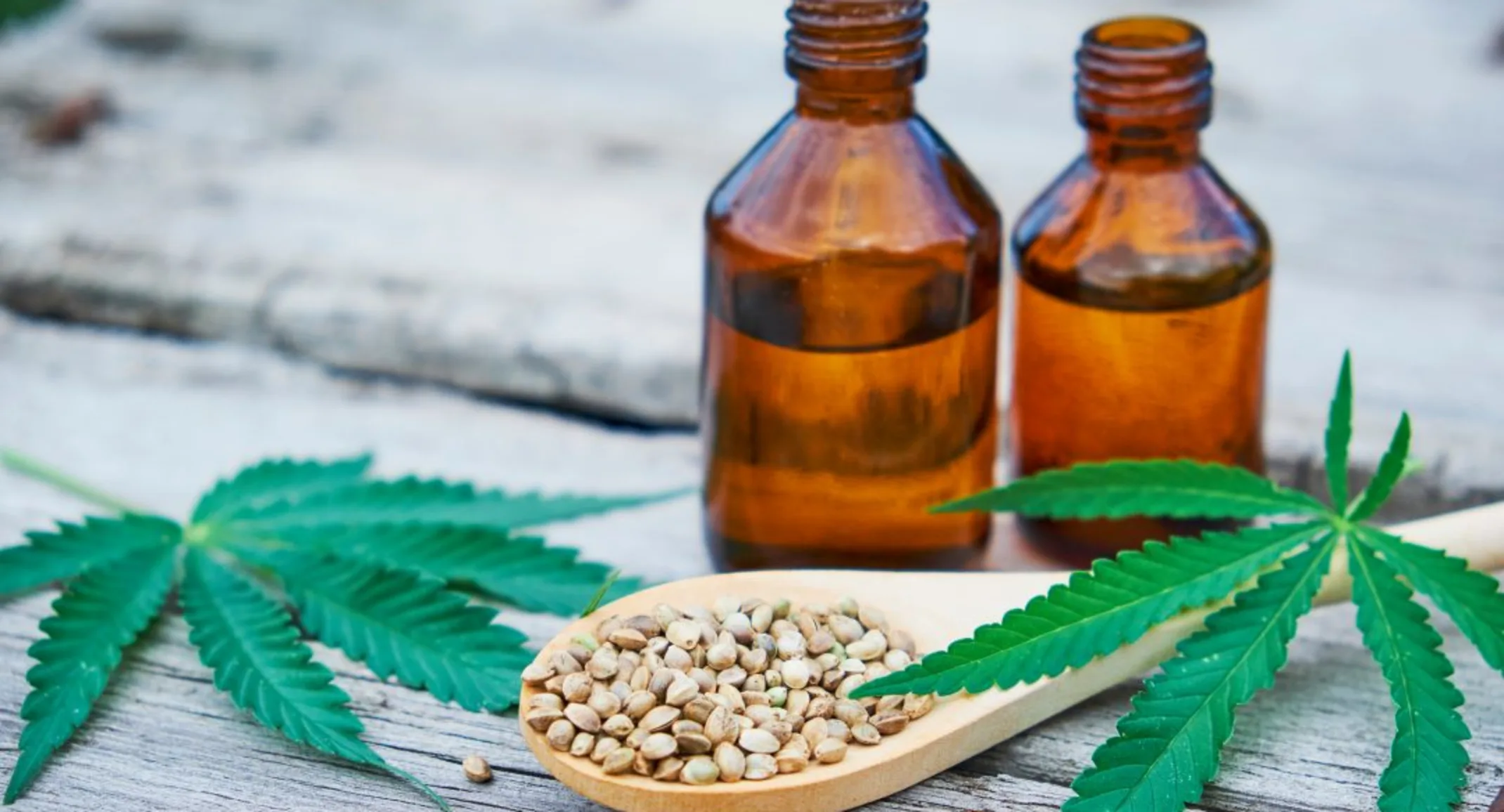 Two bottle of CDB Marijuana brown bottles next to each other next to Marijauna leaves, seeds on wooden table. 