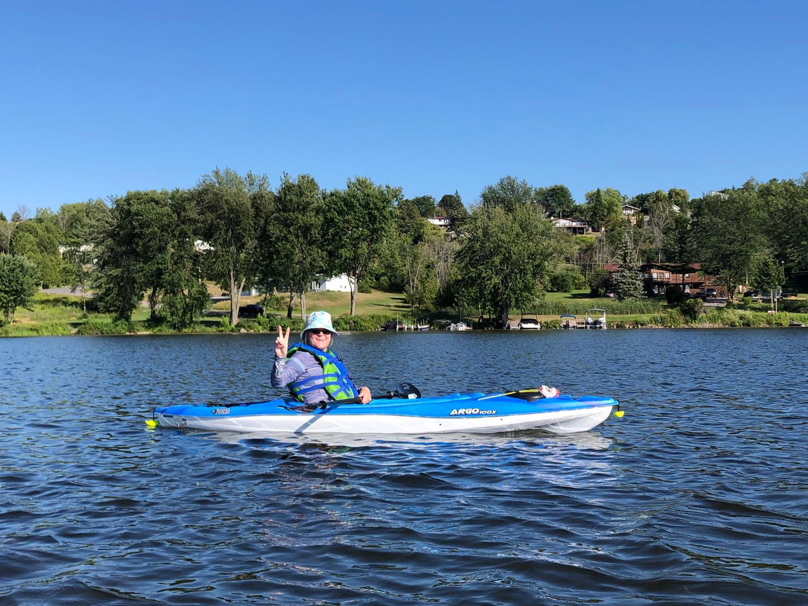 Sandra McBride smiling with hand in peace sign sitting in a kayak on a lake.