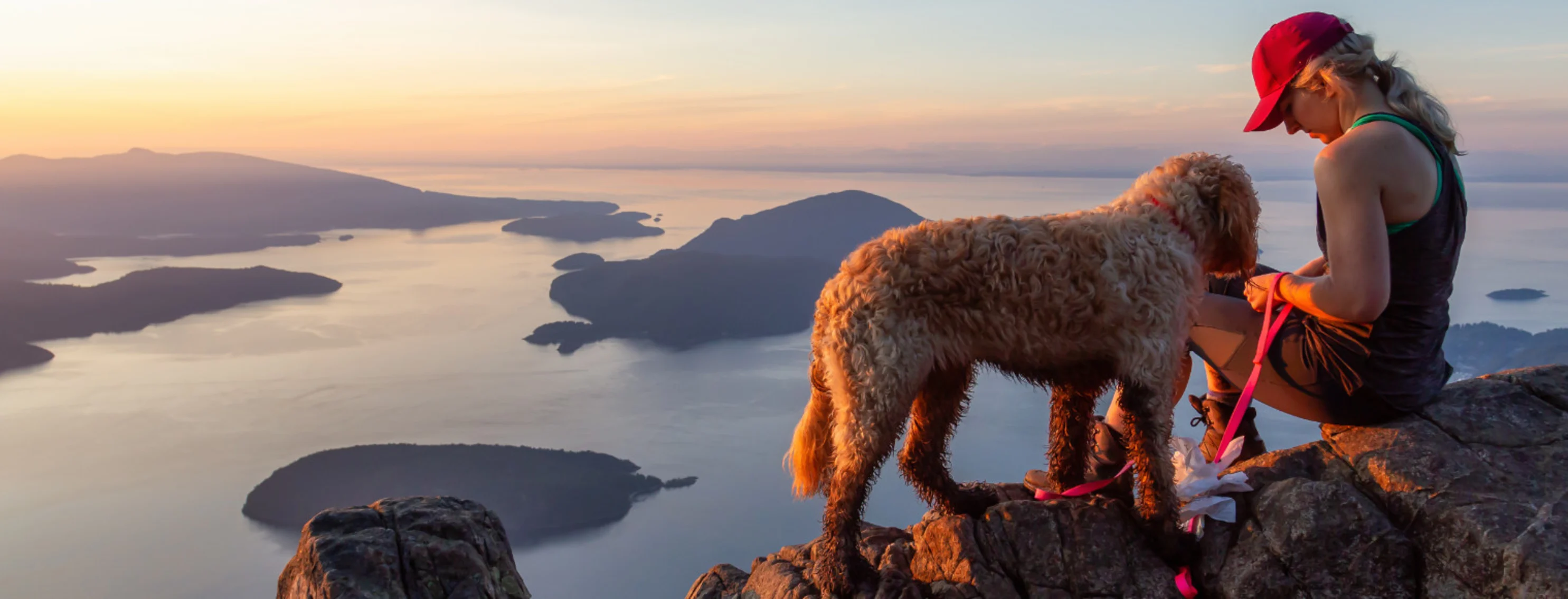 A woman and a fluffy dog sitting on a cliff overlooking water at sunset