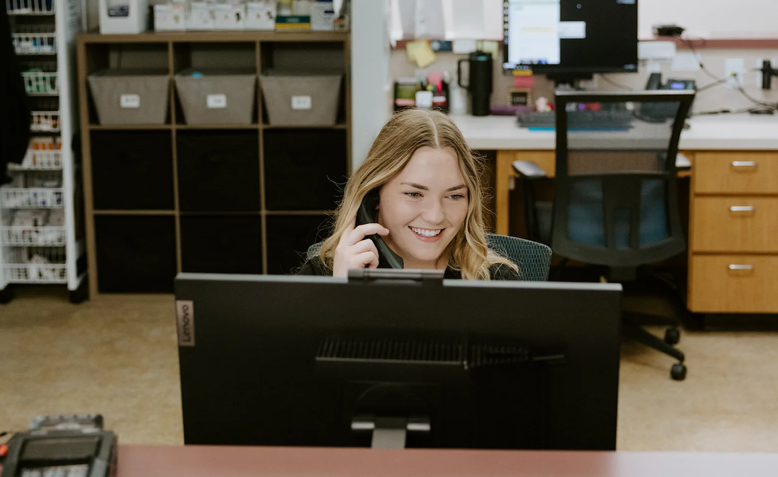 A smiling veterinary practice manager sitting at a desk behind a computer screen while talking on the phone A smiling veterinary practice manager sitting at a desk behind a computer screen while talking on the phone
