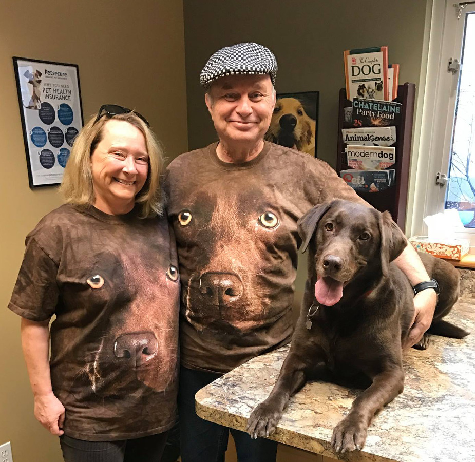 A family wearing shirts with their chocolate lab's face on them, posing with their chocolate lab.