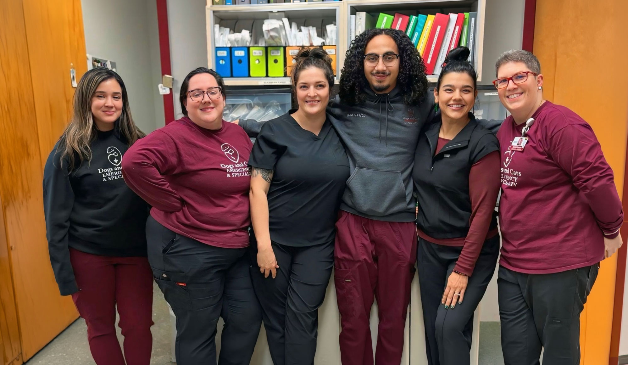 Six staff members in maroon and black scrub tops posing together in front of a supply cabinet with colorful binders