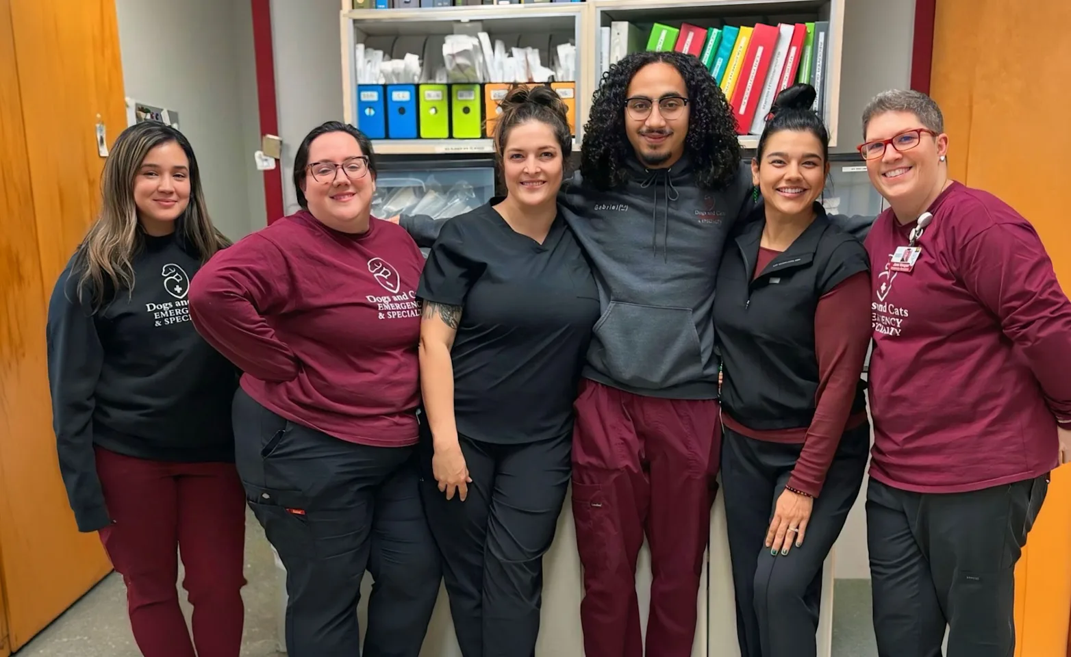 Six staff members in maroon and black scrub tops posing together in front of a supply cabinet with colorful binders Six staff members in maroon and black scrub tops posing together in front of a supply cabinet with colorful binders