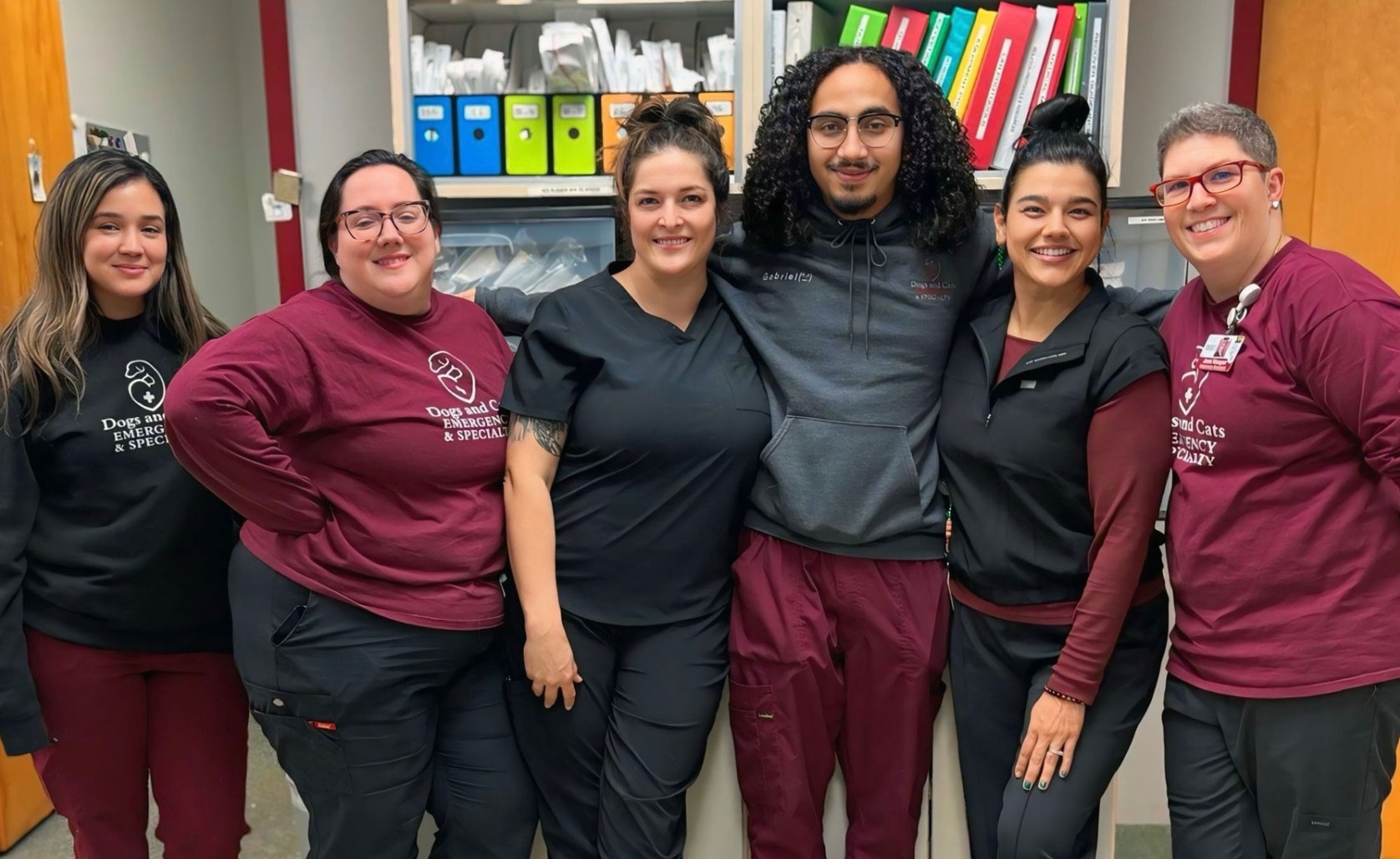 Six staff members in maroon and black scrub tops posing together in front of a supply cabinet with colorful binders