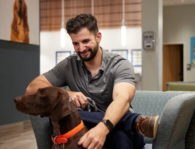 Pet parent sitting in waiting room with brown dog wearing an orange collar Pet parent sitting in waiting room with brown dog wearing an orange collar
