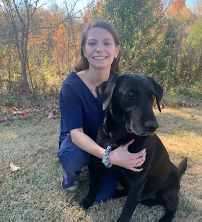 Ashley holding onto a black labrador while smiling in front of trees. Ashley holding onto a black labrador while smiling in front of trees.